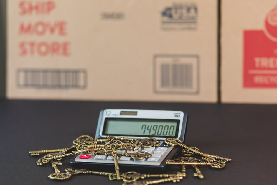 A small electronic calculator with a digital display showing the number 749,800 is placed on a dark surface. Several antique-style brass keys are scattered on top of and around the calculator, partially covering its buttons. Behind the calculator, there are large cardboard boxes stacked against a wall, with visible labels, barcodes, and printed instructions such as 'ship', 'move', and 'store' in red and black text. The boxes are in a neutral tan color and are positioned in a way that suggests they are prepared for a home relocation process. The lighting is even and neutral, highlighting the items clearly without shadows. This scene captures the process of packing and moving, involving the use of packing materials like cardboard boxes, and handling of small valuable objects like keys, which might be part of a furniture transport or home removals service offered by Man with Van Harrow. The environment appears to be an indoor storage or packing area related to moving services, reinforcing the context of relocation and logistical planning.