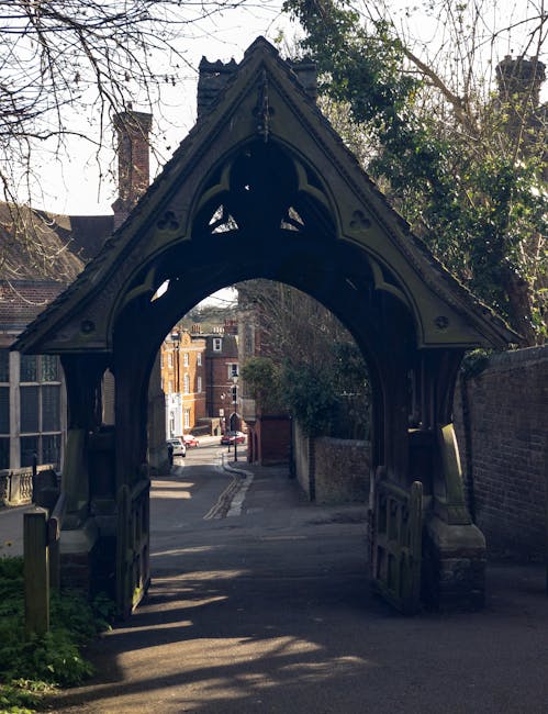 A photograph of a traditional UK brick archway with ornate wooden detailing at the top, leading into a narrow residential street. The archway appears to be an entrance to a historic property or private road, with shadows cast on the ground from nearby trees and structures. Beyond the arch, there are multi-storey brick houses with pitched roofs, along with parked cars, indicating a typical suburban neighbourhood. The scene suggests a residential area near a house or property undergoing relocation or moving preparations, with a focus on the passageway used during home removal or furniture transport. The environment is well-lit with natural daylight, and the composition captures the transition from the public street into a private property, with moving or packing activities implied by the setting outside the archway, which could be associated with services like those offered by Man with Van Harrow.