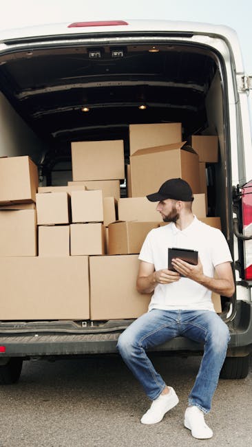 A man wearing a white t-shirt, jeans, and a black cap is sitting on the pavement next to a large moving van with its rear doors open. Inside the van, there are numerous cardboard boxes of varying sizes, some stacked on top of each other, and supported by wrapping materials. The scene suggests a home relocation process involving furniture transport and packing as part of a moving or removal service. The man appears to be reviewing information on a tablet device, likely coordinating the loading process managed by Man with Van Harrow. The background shows an outdoor environment with natural lighting, and the focus is on the organized loading of household items ready for transportation to a new property.