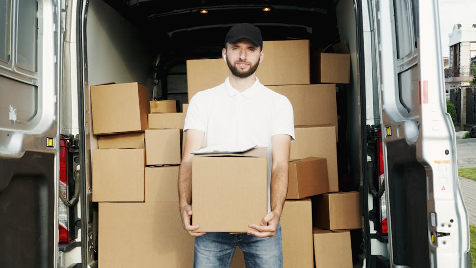 A man with a beard and wearing a white polo shirt, black cap, and blue jeans is standing inside the open rear of a white moving van, holding a cardboard box. The interior of the van is filled with numerous sealed cardboard boxes of various sizes, some stacked on each other, indicating a home relocation process. The van is parked on a paved street with residential houses visible in the background. The scene is well-lit with natural daylight, demonstrating the loading or packing phase of furniture transport and packing for a house move, as part of professional removals by Man with Van Harrow.