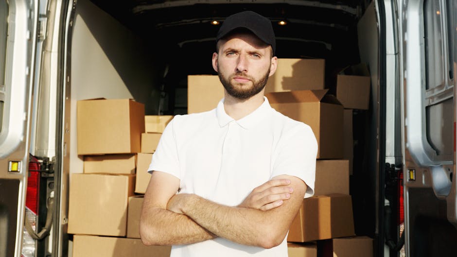 A man wearing a white t-shirt, jeans, and a black cap is sitting on the pavement next to a large moving van with its rear doors open. Inside the van, there are numerous cardboard boxes of varying sizes, some stacked on top of each other, and supported by wrapping materials. The scene suggests a home relocation process involving furniture transport and packing as part of a moving or removal service. The man appears to be reviewing information on a tablet device, likely coordinating the loading process managed by Man with Van Harrow. The background shows an outdoor environment with natural lighting, and the focus is on the organized loading of household items ready for transportation to a new property.