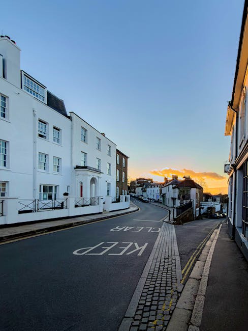 A quiet residential street in Harrow during sunset, with narrow roadways bordered by white-painted terraced houses on the left and a mixture of residential and commercial buildings on the right. The asphalt road has a 'KEEP CLEAR' sign painted on it and is partially obstructed by an unbooked van, which is used by Man with Van Harrow, a professional removals service. The sidewalk features paving stones with a drainage grate and double yellow lines along the curb. Residential windows are visible on the buildings, some with balconies and satellite dishes. The scene captures the challenges faced in house removals on narrow streets, such as vehicle access and parking constraints, illustrating the importance of experienced furniture transport and careful packing and loading processes during home relocations.