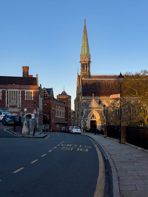 A narrow street in Harrow with residential and commercial brick buildings on the left side and a large historic church with a tall, pointed green spire on the right. The street curves slightly, with parked cars along the curb and a marked bus stop area visible on the road surface. A black lamp post is situated near the sidewalk, and trees with sparse foliage line the street. The scene is captured during daylight hours with clear blue skies. As part of a house removal or home relocation process, [COMPANY_NAME] utilises this type of environment to coordinate furniture transport and packing tasks, often involving loading procedures in tight residential streets. The setting highlights challenges with narrow street widths typical of Harrow that can affect moving logistics, and is indicative of the typical surroundings experienced during removals in such areas.