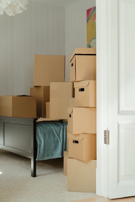 Inside a residential room showing a pile of brown cardboard moving boxes of various sizes stacked on a dark grey wooden sideboard and on the floor next to an open white door. The boxes are sealed with clear packing tape and some have built-in handles. A blue fabric item, possibly bedding or a cloth, is draped over the side of the sideboard. The room has light-colored walls with subtle vertical stripe patterns, a colorful abstract painting partially visible on the wall, and a ceiling-mounted light fixture. The scene depicts the process of home relocation and packing before furniture transport, with visible attention to safe handling and stacking of household items. A van belonging to Man with Van Harrow might be involved in the loading process outside or nearby, supporting a professional furniture removals service in Harrow.