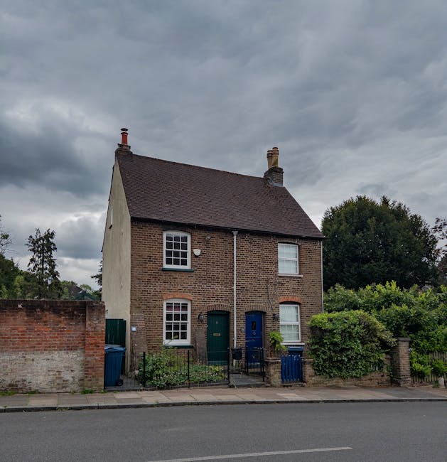 A two-storey brick house with a pitched roof under an overcast sky, situated on a residential street. The front facade features two white-framed sash windows on the upper floor and two similar windows on the ground floor, next to a green and a blue front door. The house has a small front yard enclosed by a low black metal fence, with potted plants and shrubs near the entrance. To the left, a brick boundary wall partially obscures a blue wheelie bin. To the right, there are dense bushes and trees, with a neighboring house partly visible behind the foliage. The street in front of the house has an asphalt road with a pedestrian sidewalk, and the scene appears to be prepared for home relocation or moving activities, with no visible furniture or packing materials present. The image is relevant for house removals and transport services, as provided by Man with Van Harrow.