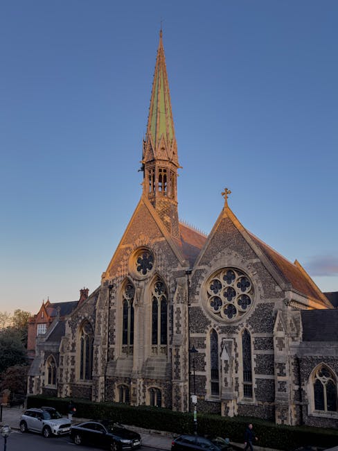 A large stone church with Gothic architectural features, including pointed arched windows, decorative tracery, and circular rose windows, set against a clear sky during daylight. The church has a tall, narrow spire with a greenish patina, topped with a small cross, and is built with a mix of lighter and darker stonework. Surrounding the church are parked cars on the street, as well as a hedge and pedestrians walking along the pavement. This image relates to house removals and relocation services, illustrating the exterior of a prominent local building in Harrow, often part of the contextual environment for furniture transport and residential moves. When conducting a home relocation or moving process, professional services like Man with Van Harrow may coordinate moving logistics around such landmarks, ensuring safe transportation of household items from inside the property to the vehicle, possibly including loading operations involving trolleys, straps, and blankets to protect furniture during the removal process.
