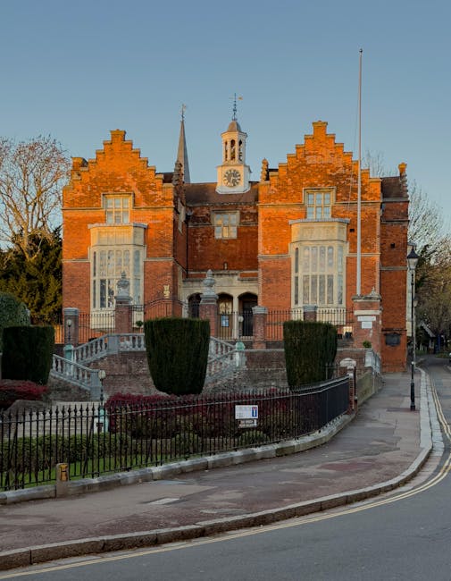 Photograph of a historic red-brick building with elaborate gabled rooflines, featuring large bay windows on the ground and upper floors, situated on a street corner with a curved sidewalk and a black metal fence enclosing a small landscaped garden. The garden contains well-trimmed bushes and a pathway leading to a front entrance with steps, surrounded by concrete ramp access, and a white sign on the fence. In the background, there are tall trees and a clear blue sky, with a lamp post near the corner of the street. The scene depicts a typical urban area suitable for home relocation or moving services. The image is clear and well-lit, capturing the architectural details and street environment associated with property moving logistics, aligning with services offered by Man with Van Harrow.