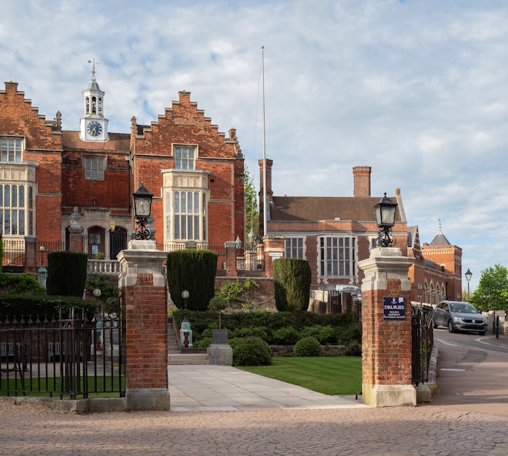 Photograph of a historic red-brick building with elaborate gabled rooflines, featuring large bay windows on the ground and upper floors, situated on a street corner with a curved sidewalk and a black metal fence enclosing a small landscaped garden. The garden contains well-trimmed bushes and a pathway leading to a front entrance with steps, surrounded by concrete ramp access, and a white sign on the fence. In the background, there are tall trees and a clear blue sky, with a lamp post near the corner of the street. The scene depicts a typical urban area suitable for home relocation or moving services. The image is clear and well-lit, capturing the architectural details and street environment associated with property moving logistics, aligning with services offered by Man with Van Harrow.