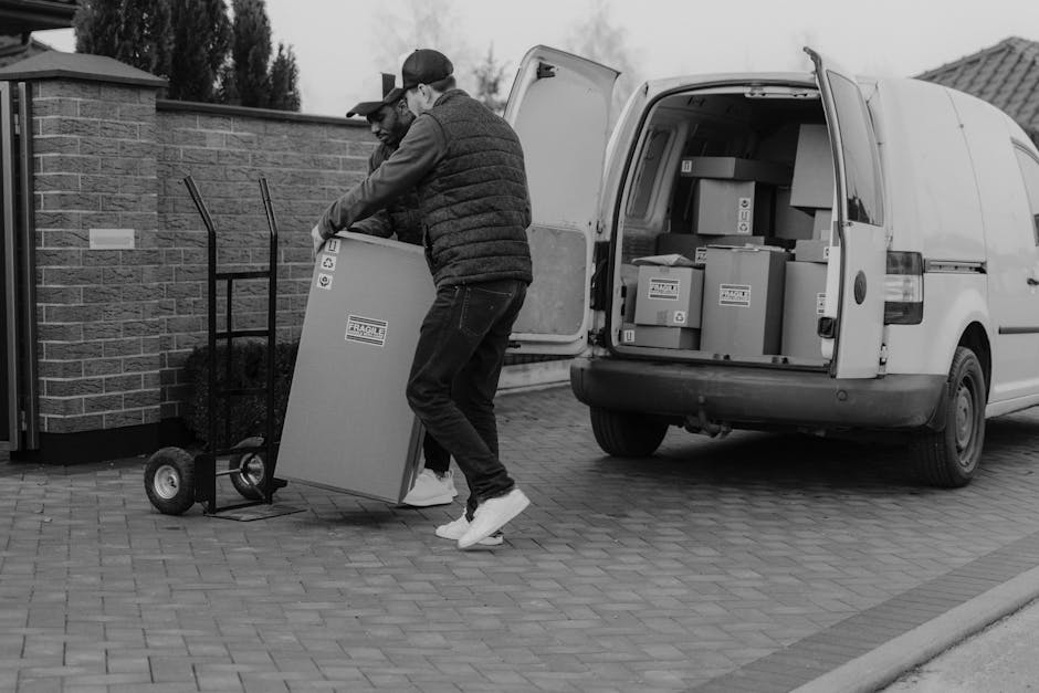 Two movers from Man with Van Harrow are in the process of loading a large, rectangular cardboard box with a fragile label onto a flat dollie. One mover, wearing a baseball cap and dark clothing, is he