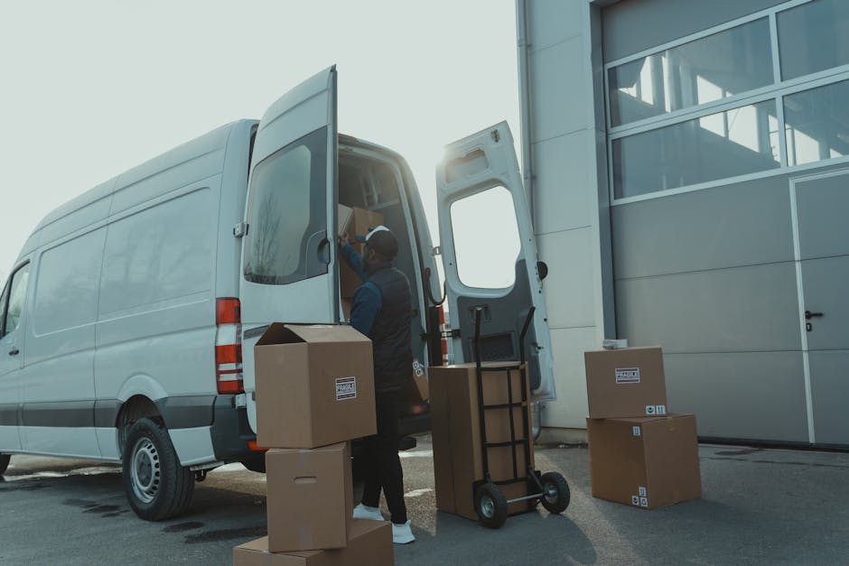 Two movers from Man with Van Harrow are in the process of loading a large, rectangular cardboard box with a fragile label onto a flat dollie. One mover, wearing a baseball cap and dark clothing, is he