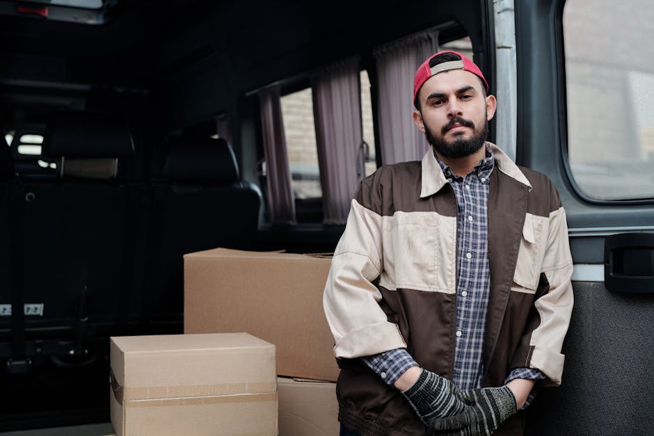 A man with a beard and wearing a grey and beige jacket, blue checkered shirt, and red cap stands inside the open rear of a van, which is parked outside a building during daytime. He is positioned beside several neatly packed cardboard boxes, some wrapped with packing tape, ready for a home relocation or furniture transport. The interior of the van is visible with black seats and a partially visible window with curtains. The scene captures the loading process associated with house removals, with the man prepared to assist with packing and moving tasks. The environment is well-lit, emphasizing the professional effort involved in packing and transporting household items with a van as part of the moving services provided by Man with Van Harrow, aligned with the context of urgent same-day relocation tips.