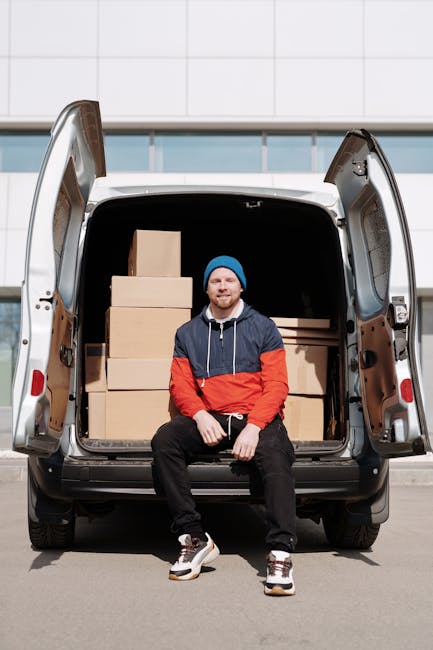A man with a beard wearing a black cap and a white polo shirt stands inside the back of a moving van filled with uniformly sized cardboard moving boxes. The boxes are stacked towards the front and sides of the van's interior, some wrapped in plastic or packing materials. The man has his arms crossed and appears to be observing the loading process, which takes place outdoors on a paved surface. The van is open at the rear, revealing the boxes and part of the driveway or yard. This scene depicts the logistics of a home relocation, with the focus on furniture transport, packing, and loading during a house move, highlighting the professional services provided by Man with Van Harrow for removals and moving logistics.
