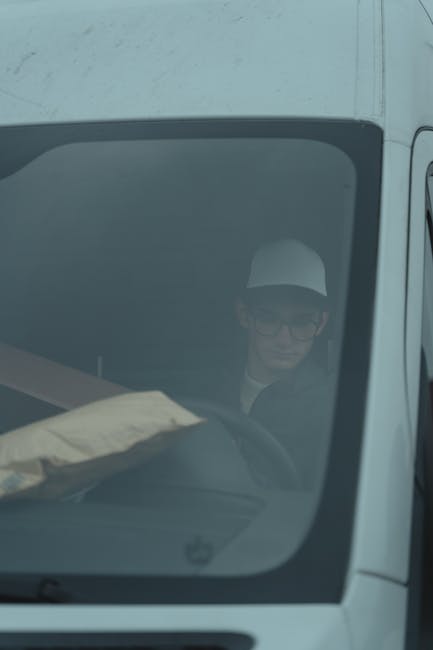 A person wearing glasses and a white cap is seated inside a white van, visible through the front windshield. The individual appears to be focused on the vehicle's interior, possibly preparing for or during a home relocation process. The van's dashboard and steering wheel are partially visible, with some packing materials, such as a cardboard box wrapped in plastic, placed on the dashboard. The background outside the vehicle shows a glimpse of a building or street, indicating the vehicle is parked in an urban or residential area. The scene suggests a furniture transport or removal operation taken by Man with Van Harrow as part of their home removals services, with attention to careful handling and space management during the loading process.