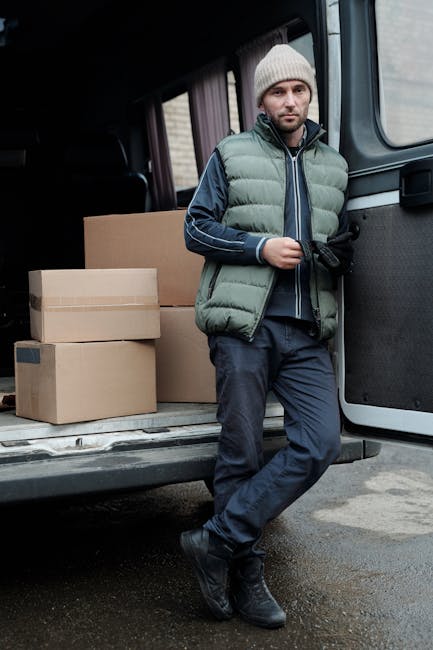 Two men are engaged in a home relocation process, inside a paved driveway of a residential property with a brick wall and hedges in the background. One man, wearing a black quilted vest and dark jeans, is lifting or stabilising a large cardboard box, while the other, dressed in a blue and black jacket with white trainers, is assisting by supporting the box from the opposite side. The open rear doors of a white delivery van reveal numerous packed cardboard boxes organized inside, indicating a packing and moving operation. Nearby, a two-wheeled hand truck with a black handle and wheels stands on the driveway, ready for furniture transport or loading tasks. The scene suggests a professional removals service, such as Man with Van Harrow, handling a house move with attention to secure packing and efficient loading, suitable for tight access situations as discussed in the Wealdstone man with van insider tips for tight access, HARROW.